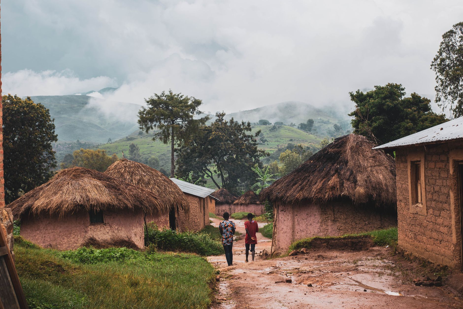 Two men walking through a rural village with thatched roof huts and lush greenery.