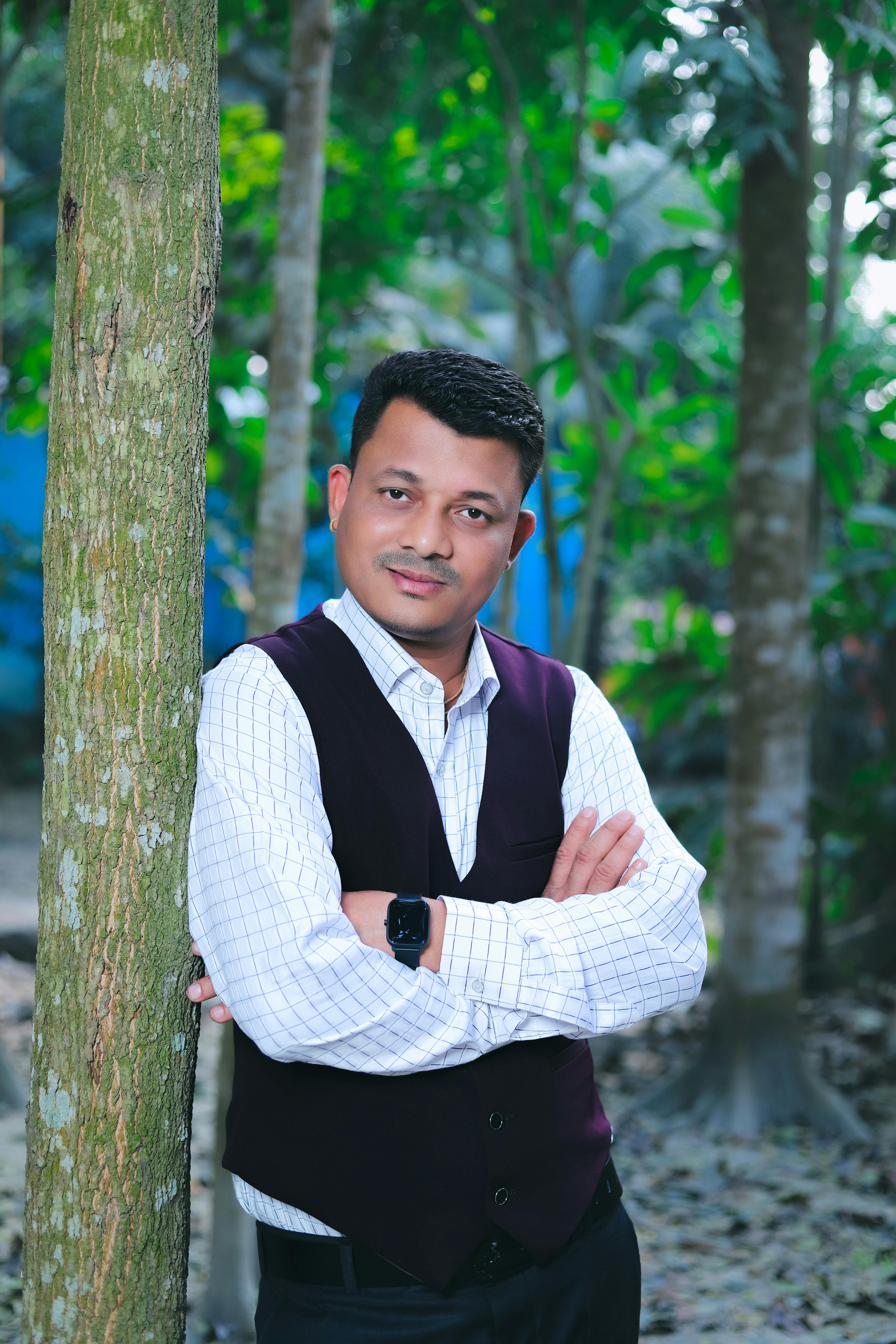 A confident man in a vest posing against a tree in a lush Bangladeshi setting.