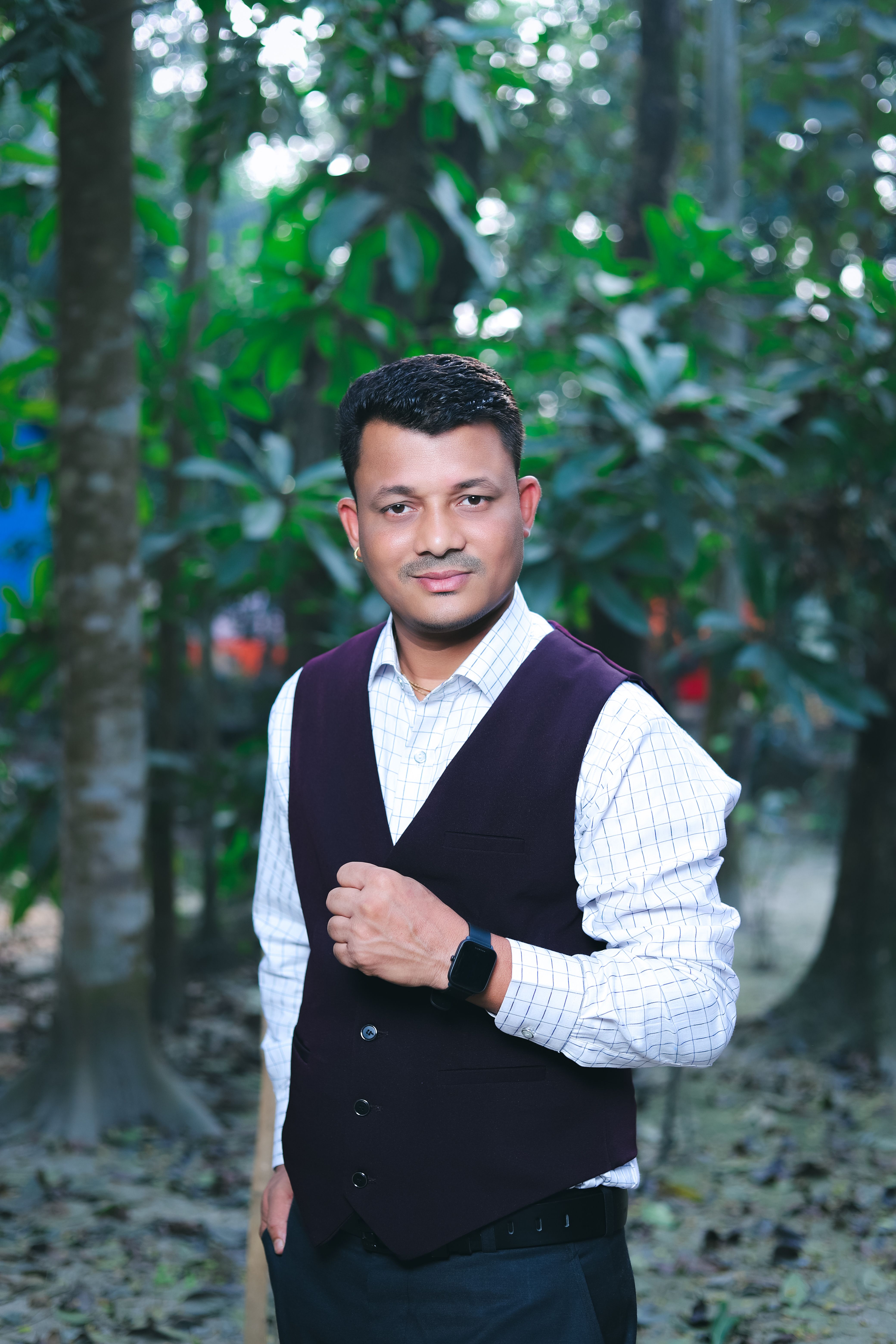 A confident man in formal attire poses amidst lush greenery.