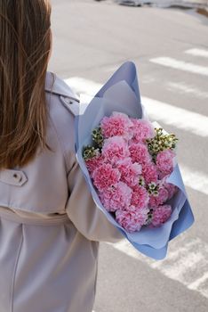 A woman in a beige coat holds a bouquet of pink carnations crossing a city street.