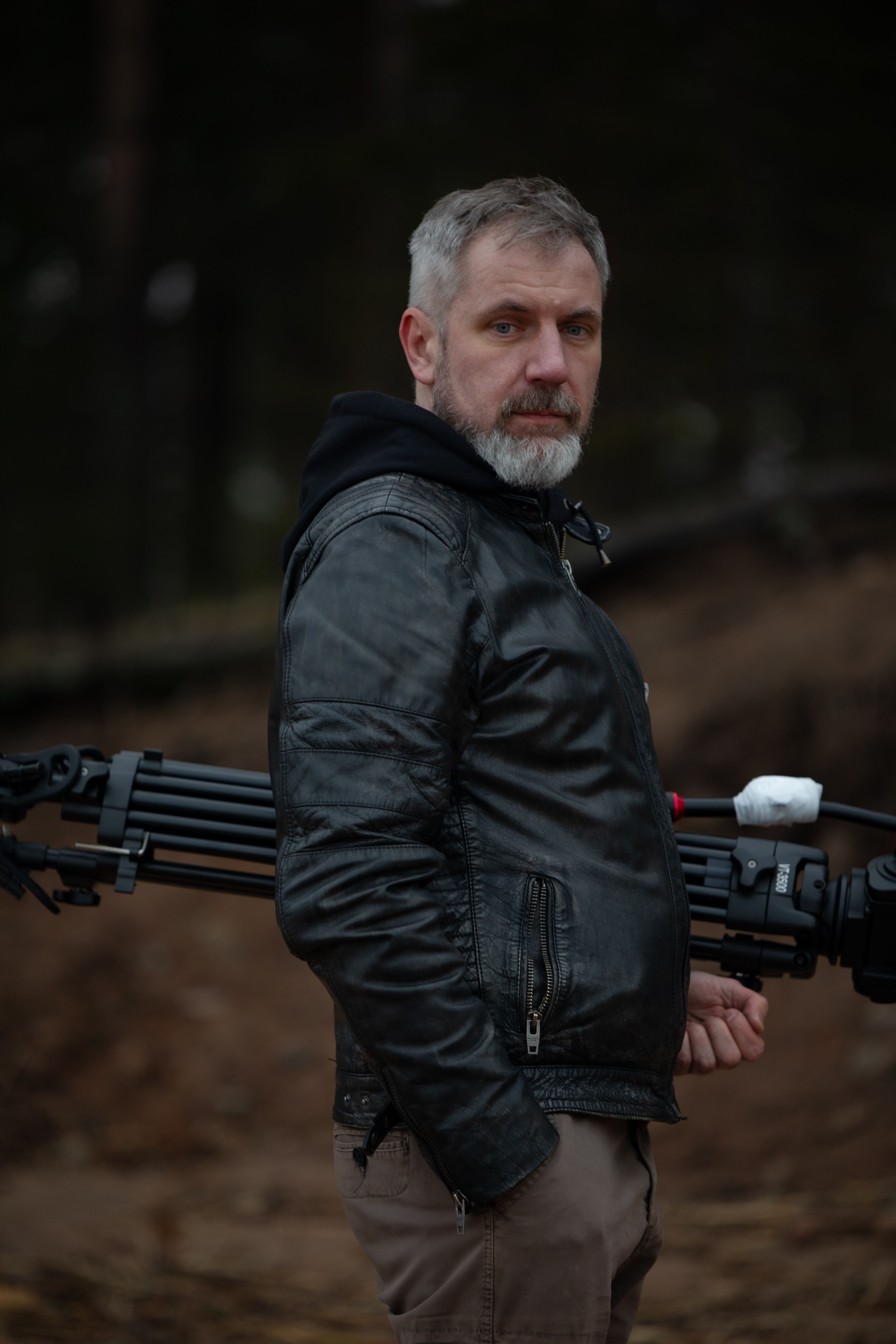 A rugged man in a leather jacket holding a photography tripod outdoors.