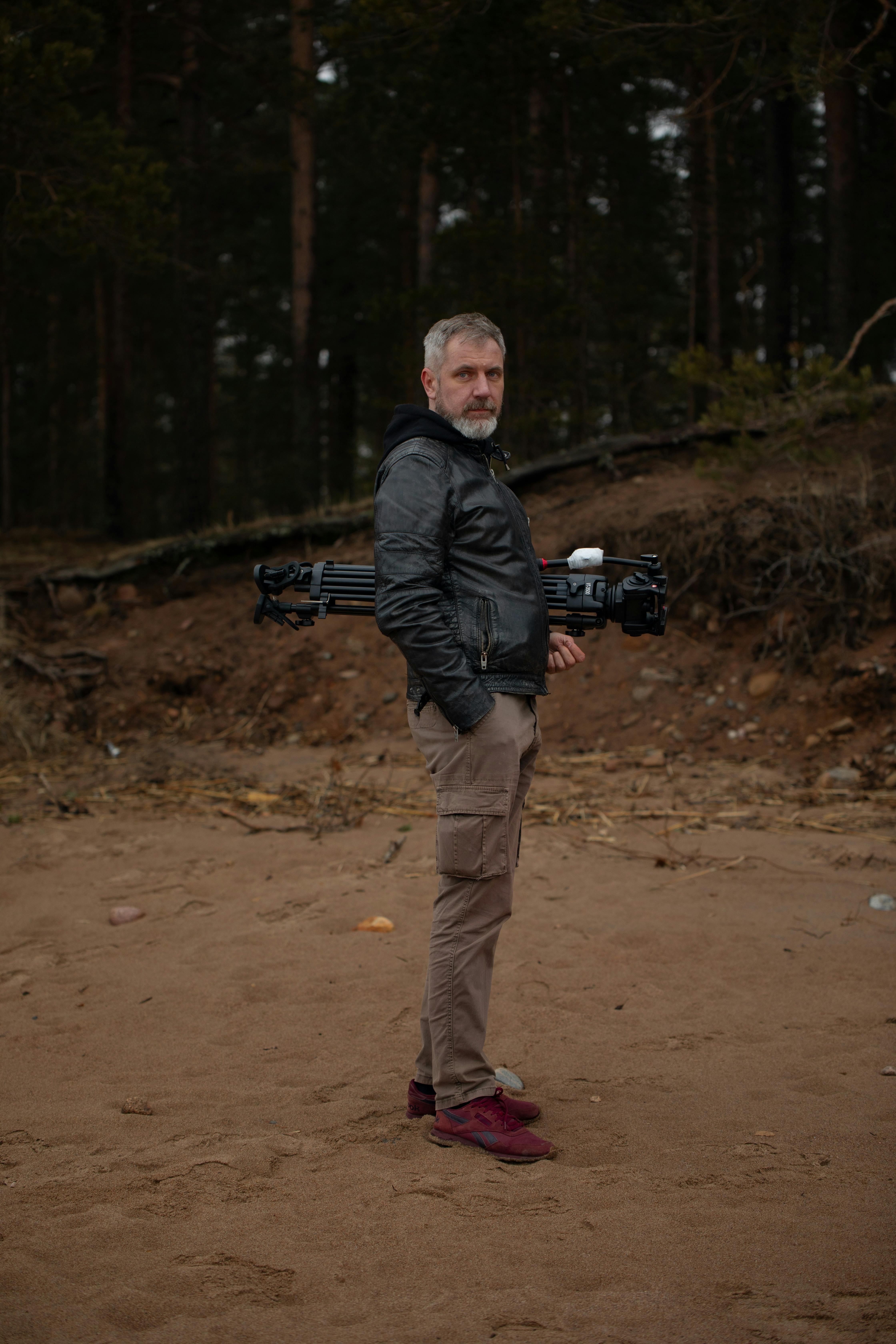 Older man holding a tripod, wearing a leather jacket, standing in forest clearing.