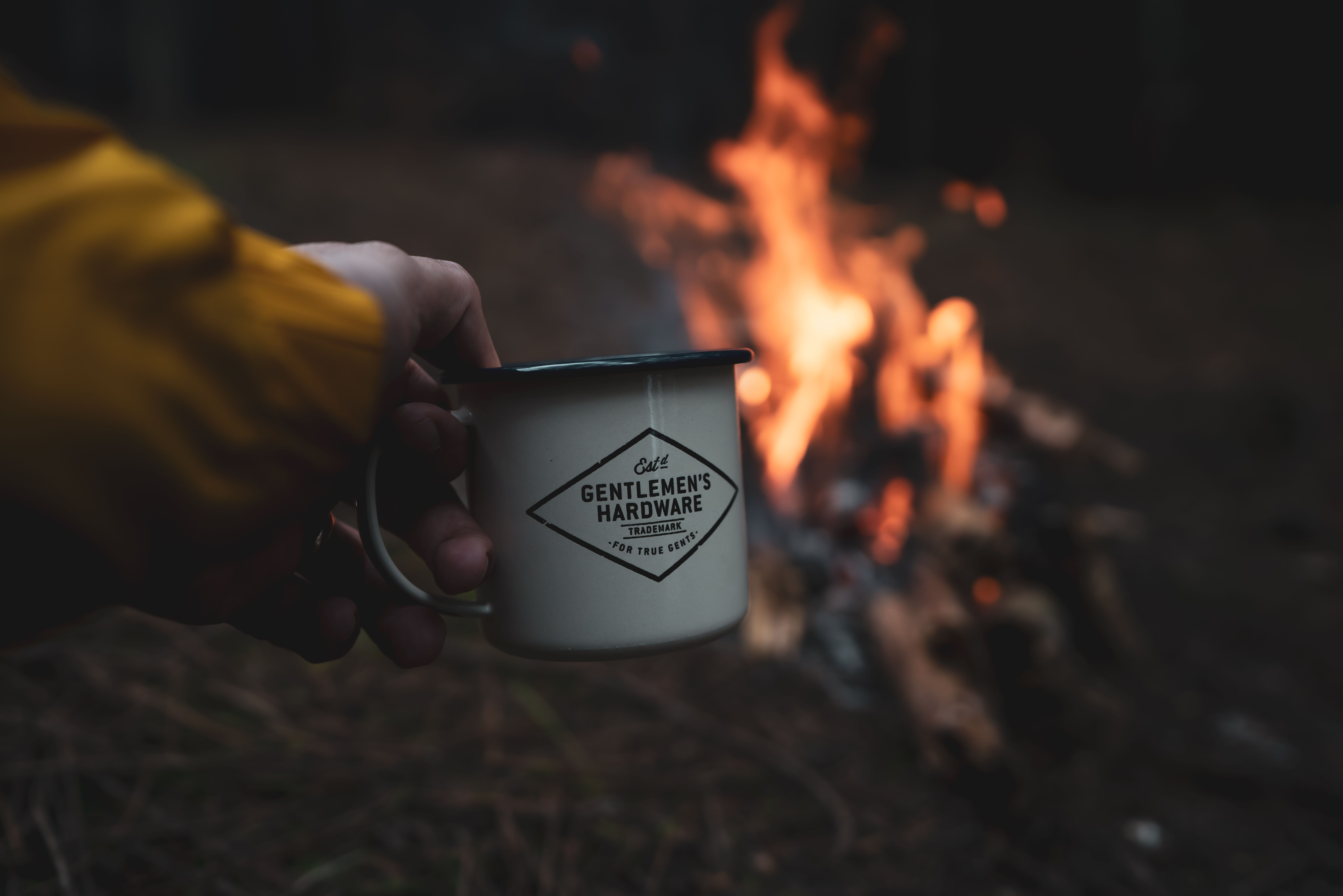 A hand holding a mug in front of a warm campfire, creating a cozy outdoor vibe.
