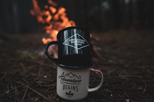 A mug with the words adventure begins in front of a campfire