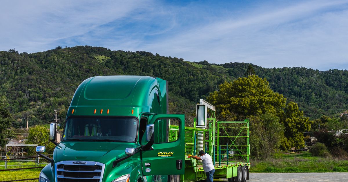 A vivid green truck with a trailer parked in a scenic landscape with hills and greenery.