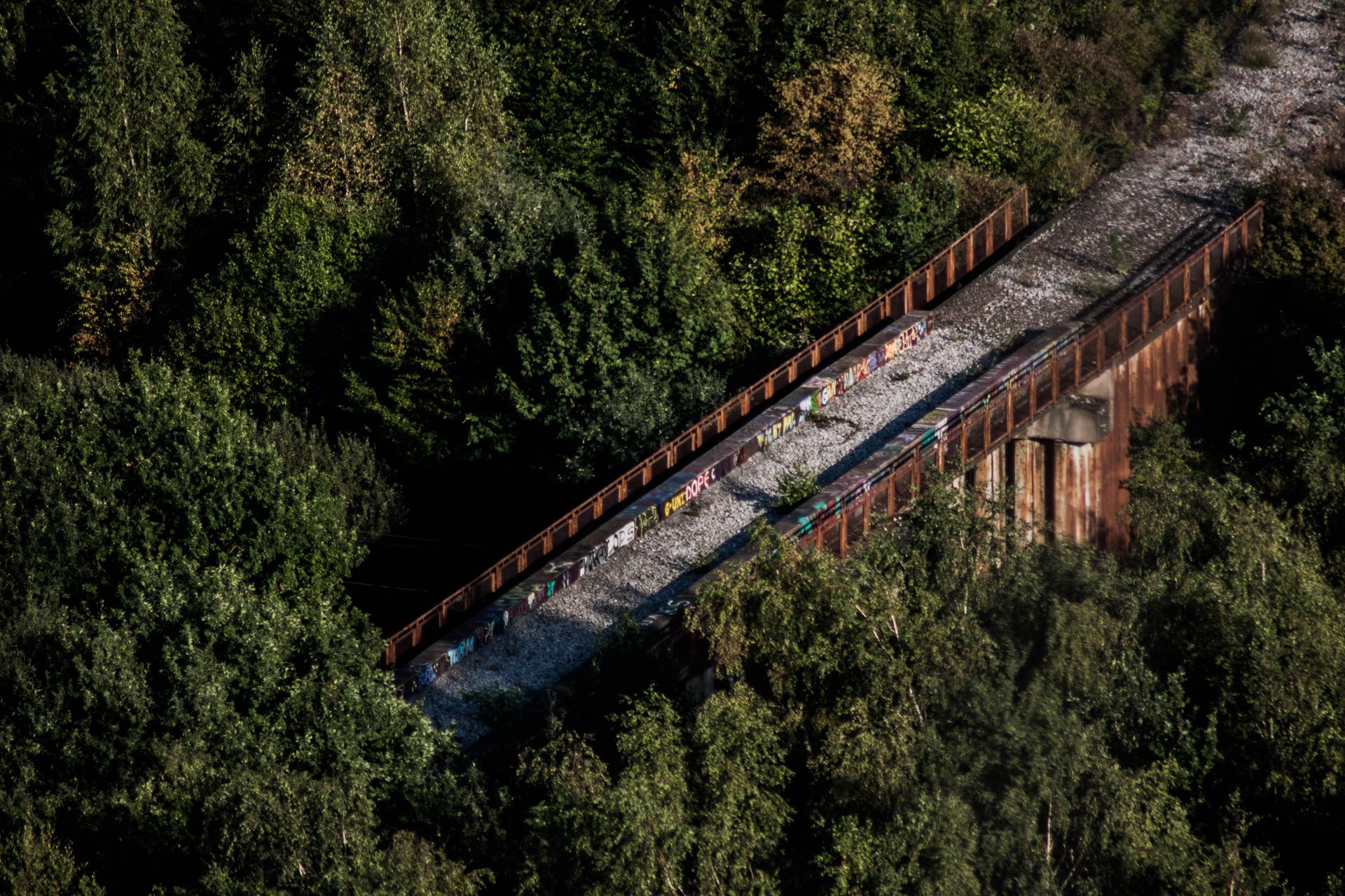 Aerial shot of an overgrown, graffiti-covered abandoned railway bridge surrounded by dense forest.