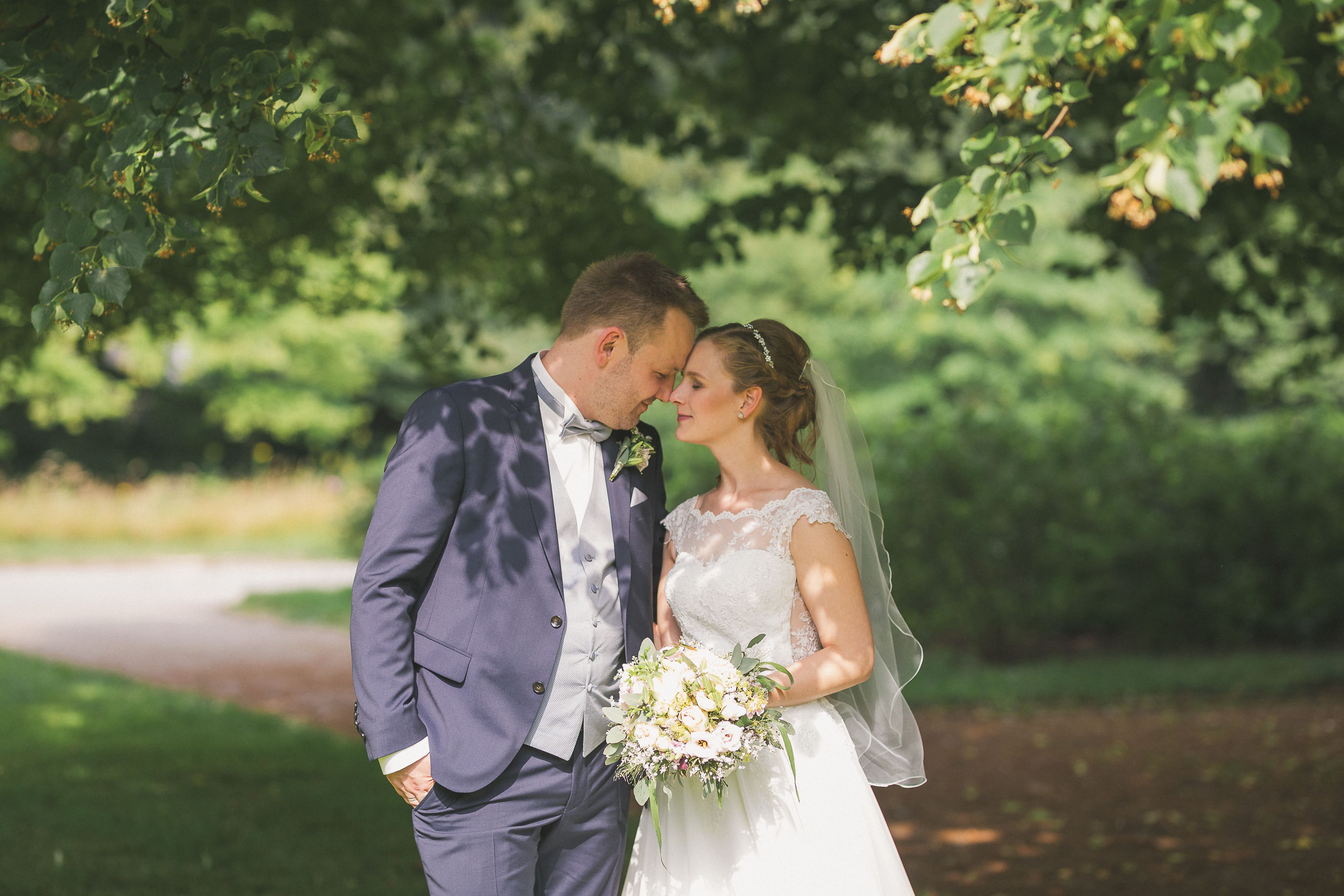 Bride and groom embracing under trees in a sunlit garden, sharing a tender moment during their wedding day.