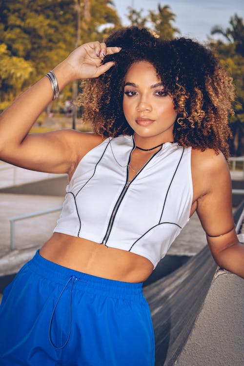 A woman with curly hair posing in a white crop top and blue pants