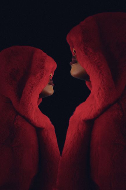 Artistic photo of two red hooded figures facing each other against a dark backdrop.