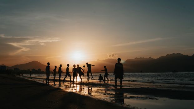 A group of people playing football on a sandy beach during sunset, creating stunning silhouettes against the ocean backdrop.
