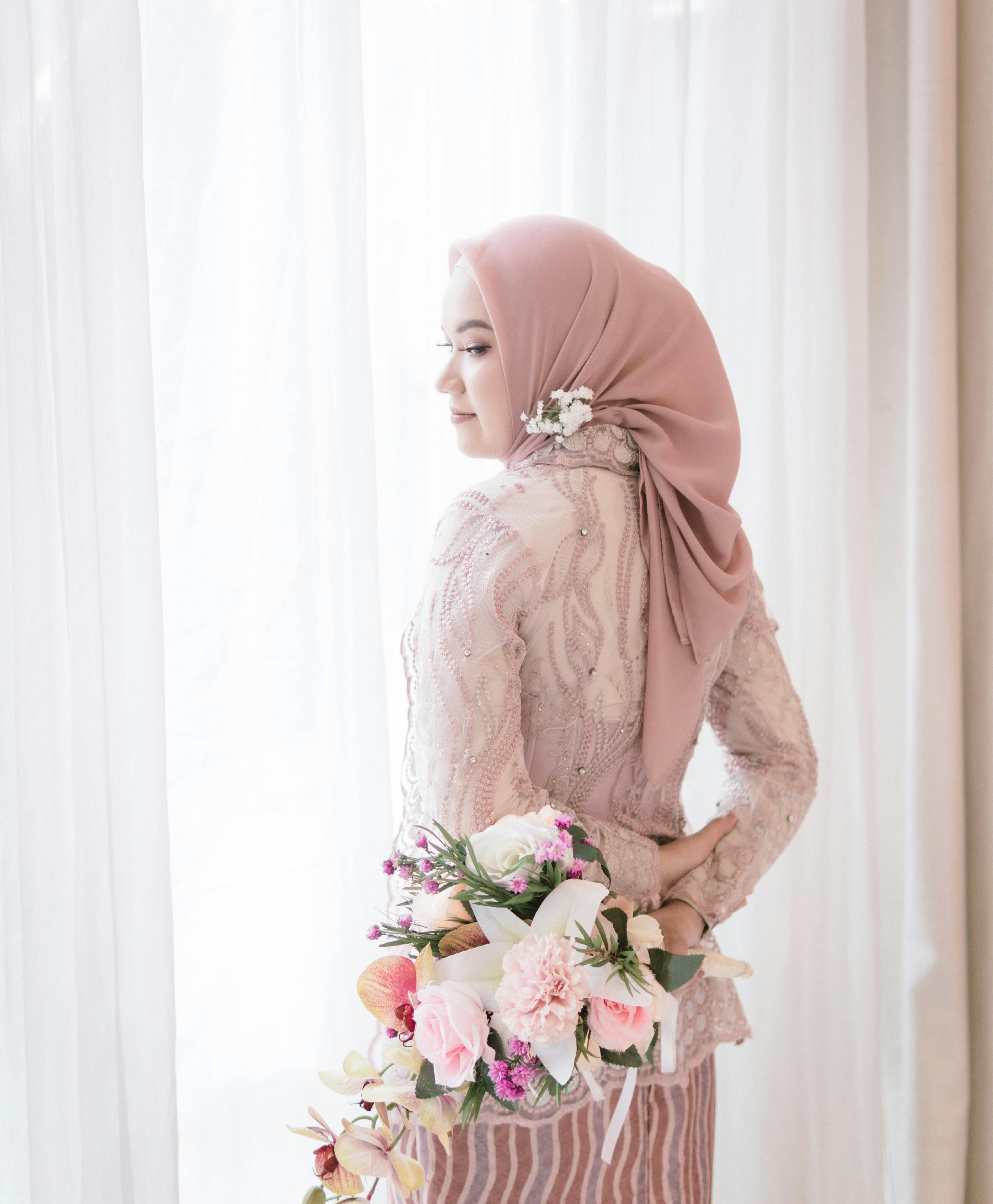A young woman in a pink hijab elegantly holds a bouquet of flowers indoors, exuding style and grace.
