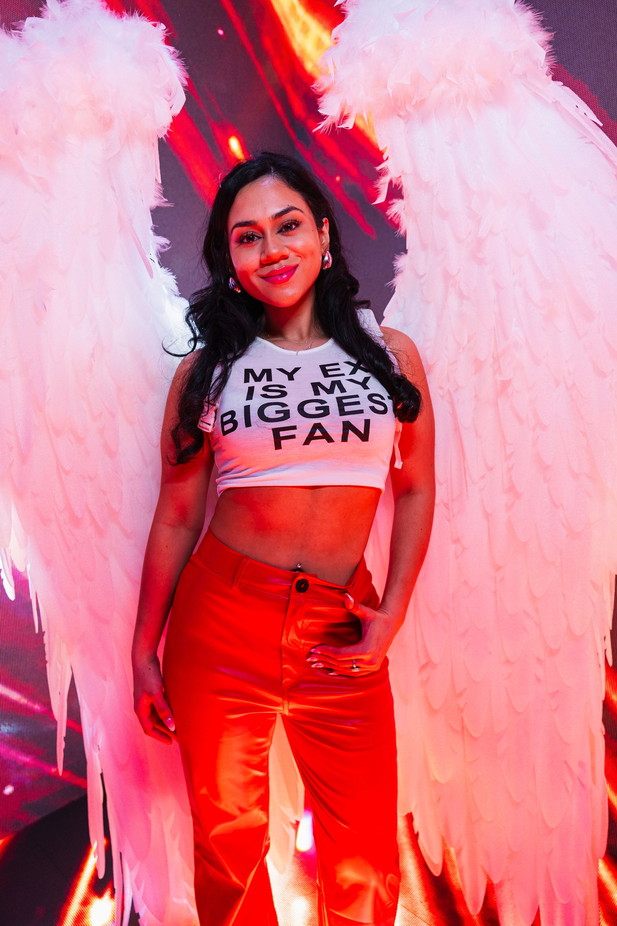 Woman posing with angel wings in a nightclub, wearing red pants and a tank top.