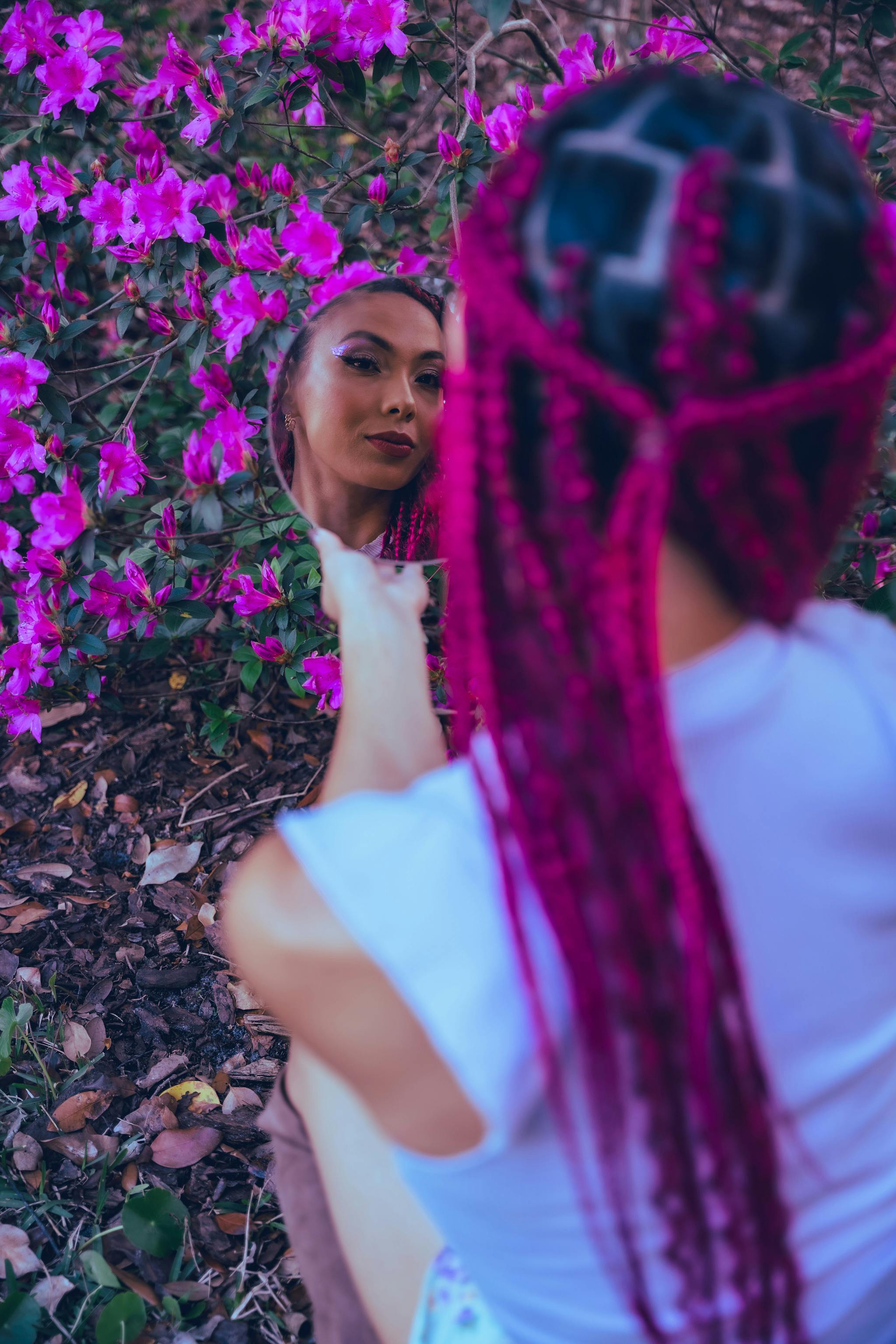 Woman with pink braids admiring her reflection in nature surrounded by flowers.