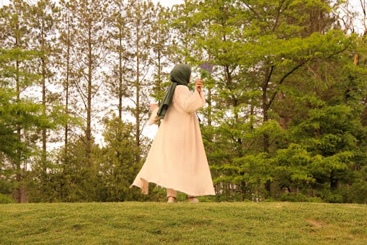 A serene woman in a hijab holding flowers, walking in a lush green park during spring.