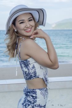 Charming woman in a hat and swimsuit smiles on a breezy seaside day.
