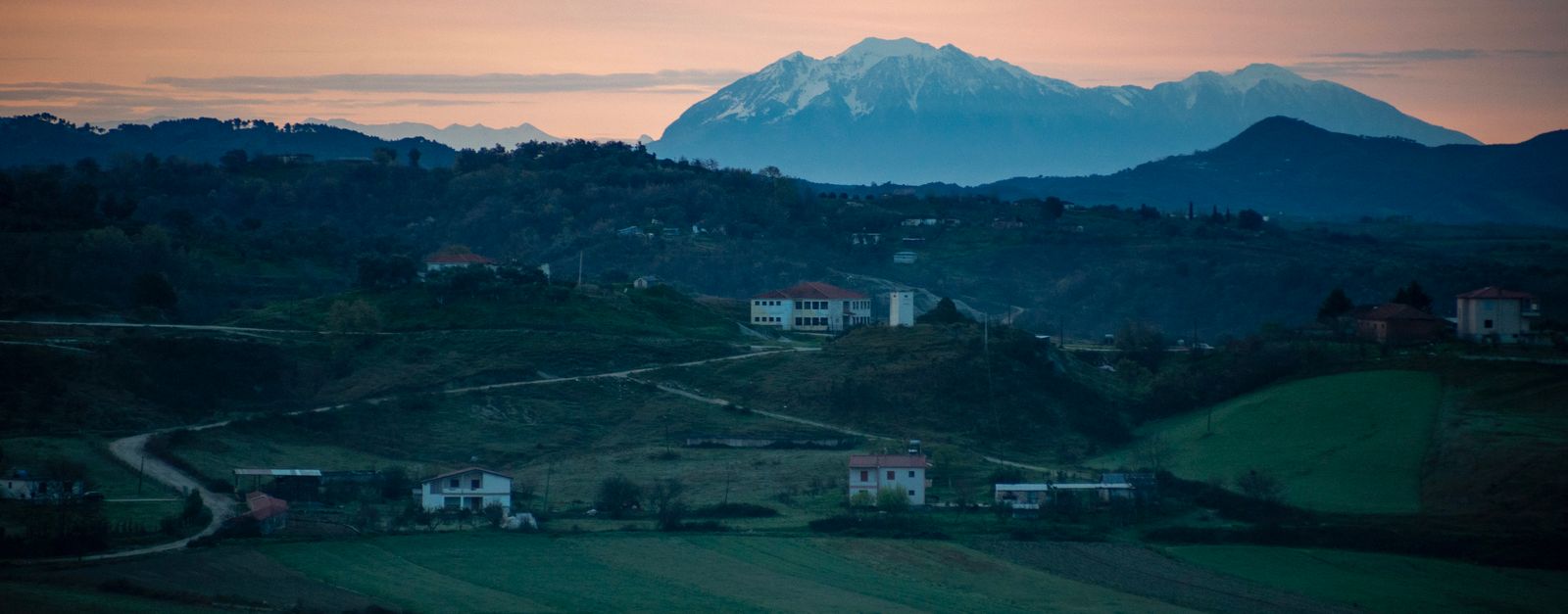 A tranquil rural scene in Albania, showcasing rolling hills and mountains under a sunset sky.
