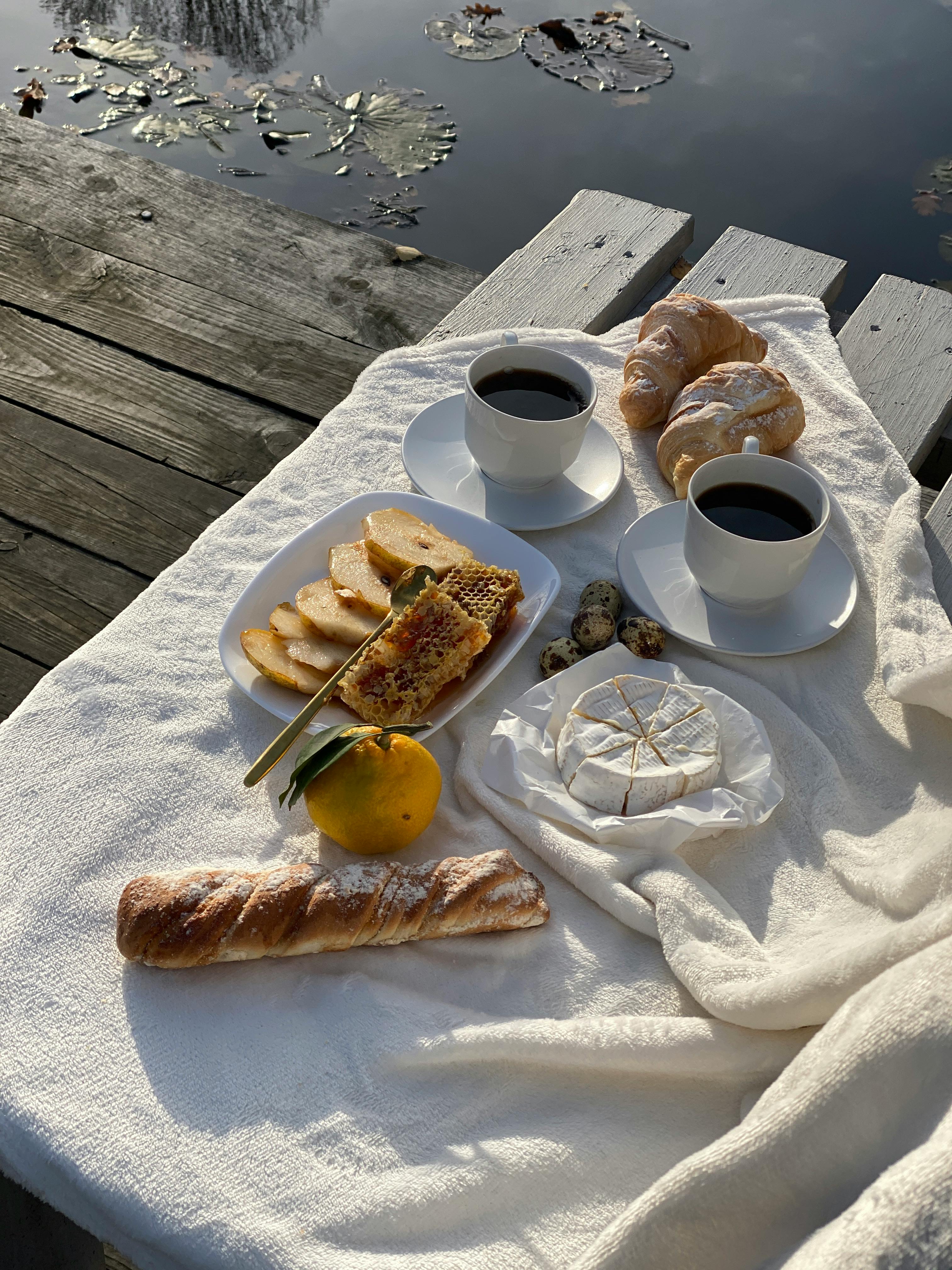 A cozy, elegant brunch setup on a pier with cheese, croissants, and coffee by the water.
