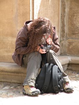 A woman with long hair checks her camera while seated outdoors, next to a stone wall.
