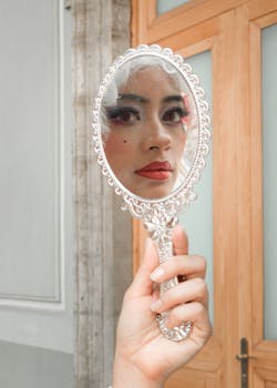 Close-up reflection of a young woman wearing makeup in an ornate handheld mirror indoors.