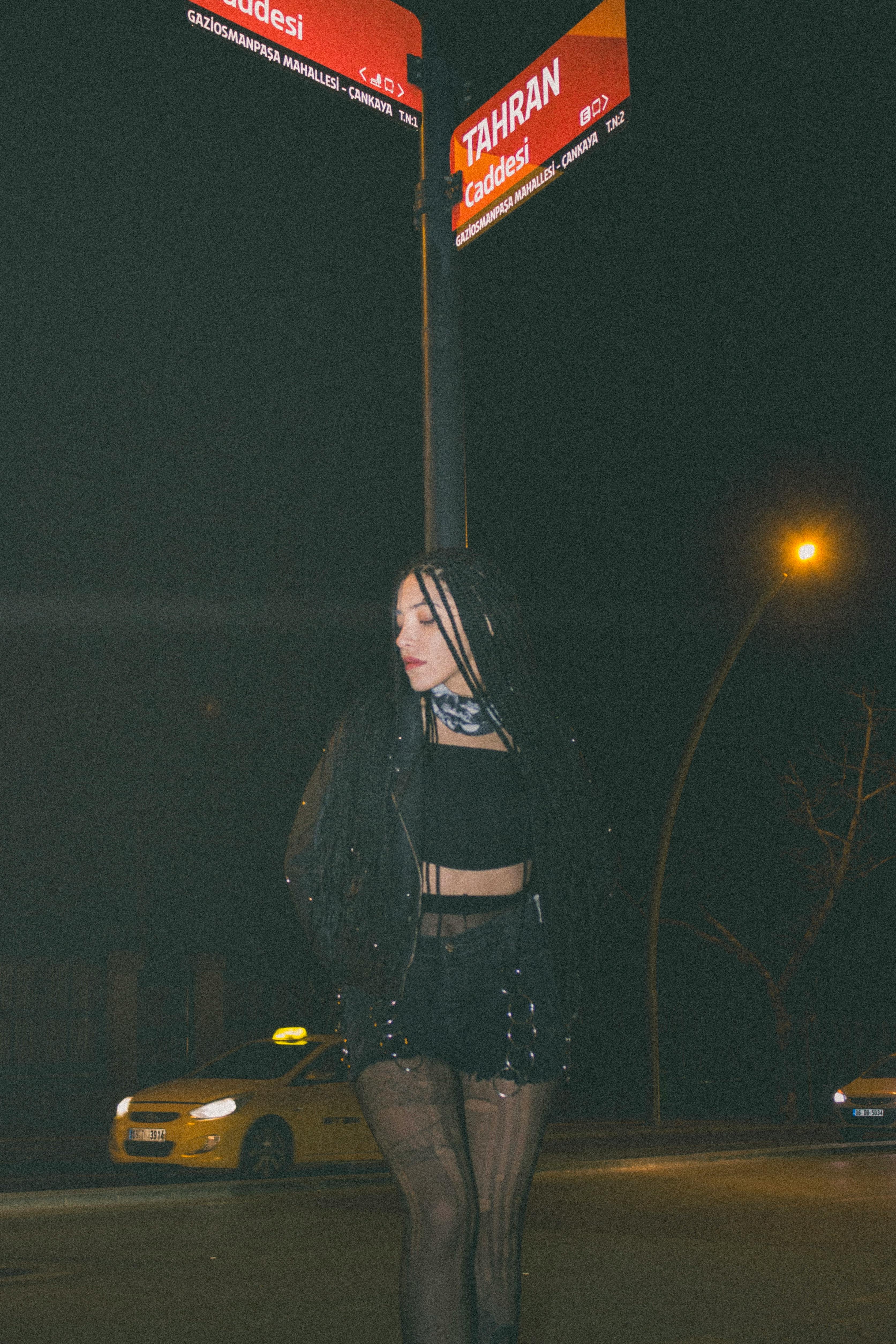 A woman stands under a street sign in Ankara at night, dressed in black attire.