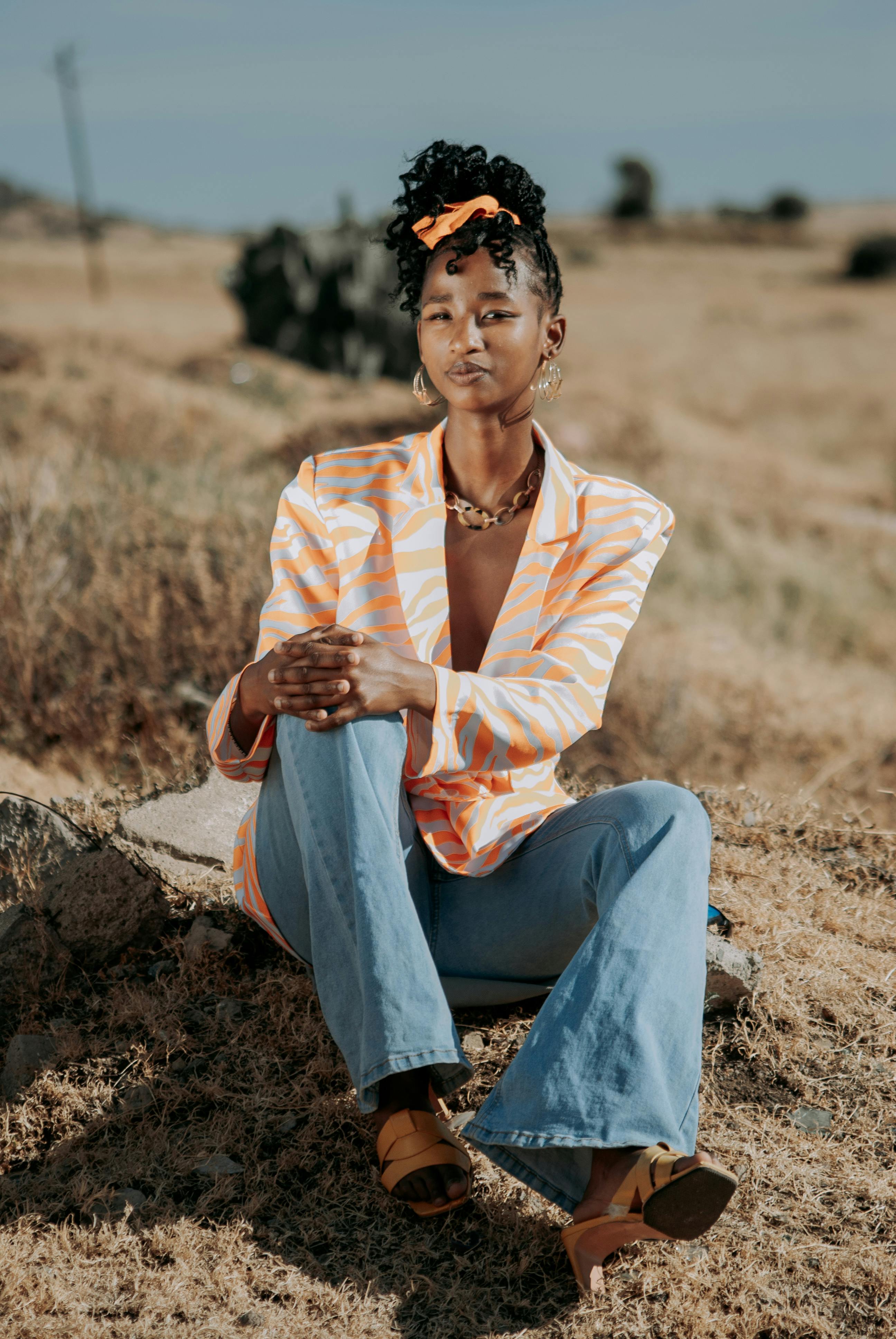 Stylish woman in vibrant suit jacket and jeans sitting outdoors in a rural area.