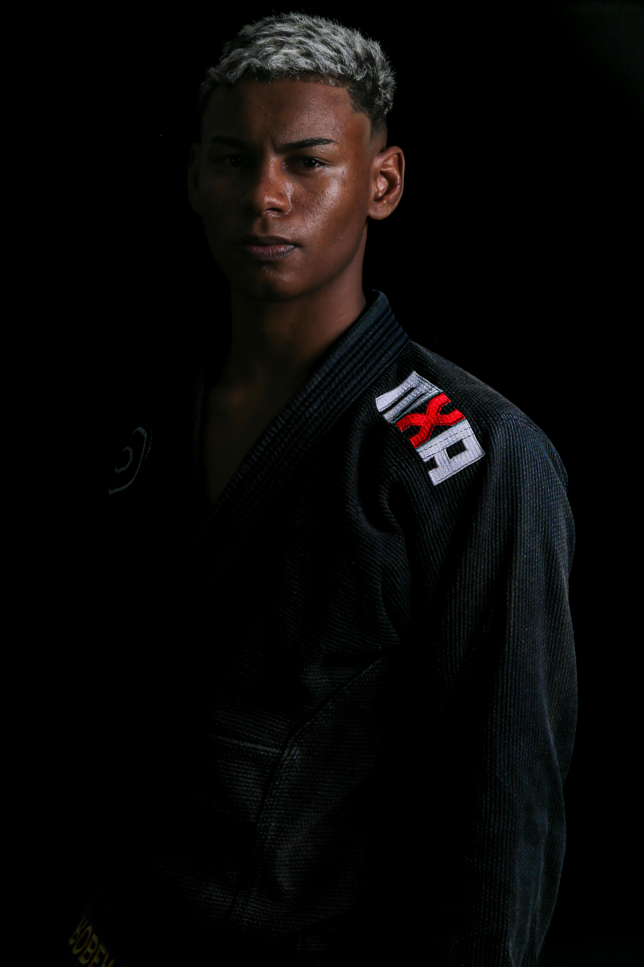 Confident martial artist in black gi with embroidered logo on black background.