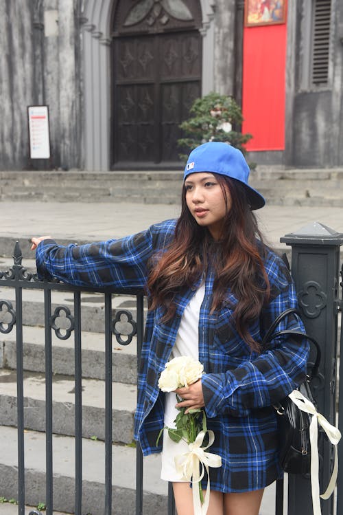 A woman in a plaid shirt and blue hat is leaning against a fence
