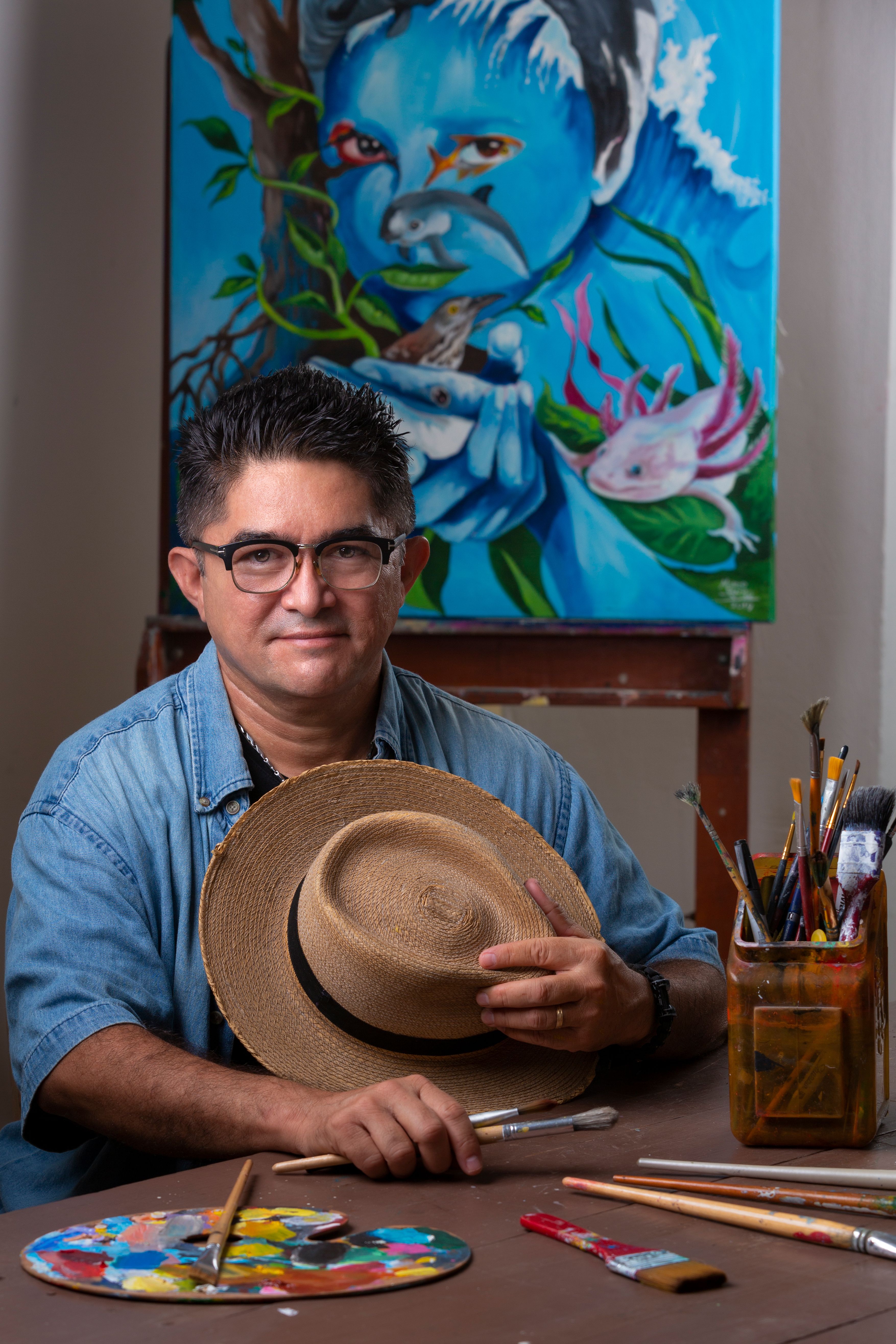 An artist poses with a hat, brushes, and a blue-themed painting in a studio.