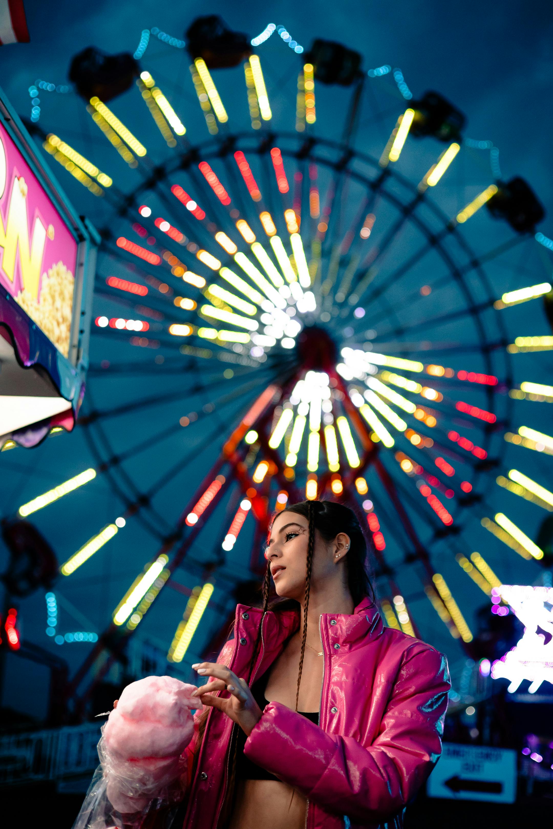 A young woman in a pink jacket enjoys cotton candy at a lively amusement park night scene.