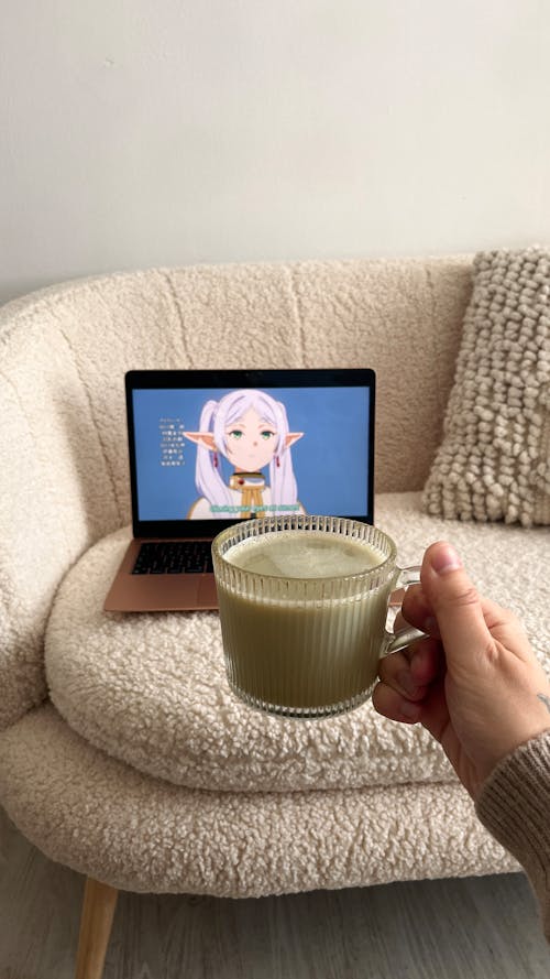 A hand holding a green matcha latte over a MacBook Air playing "Frieren: Beyond The Journey's End" 