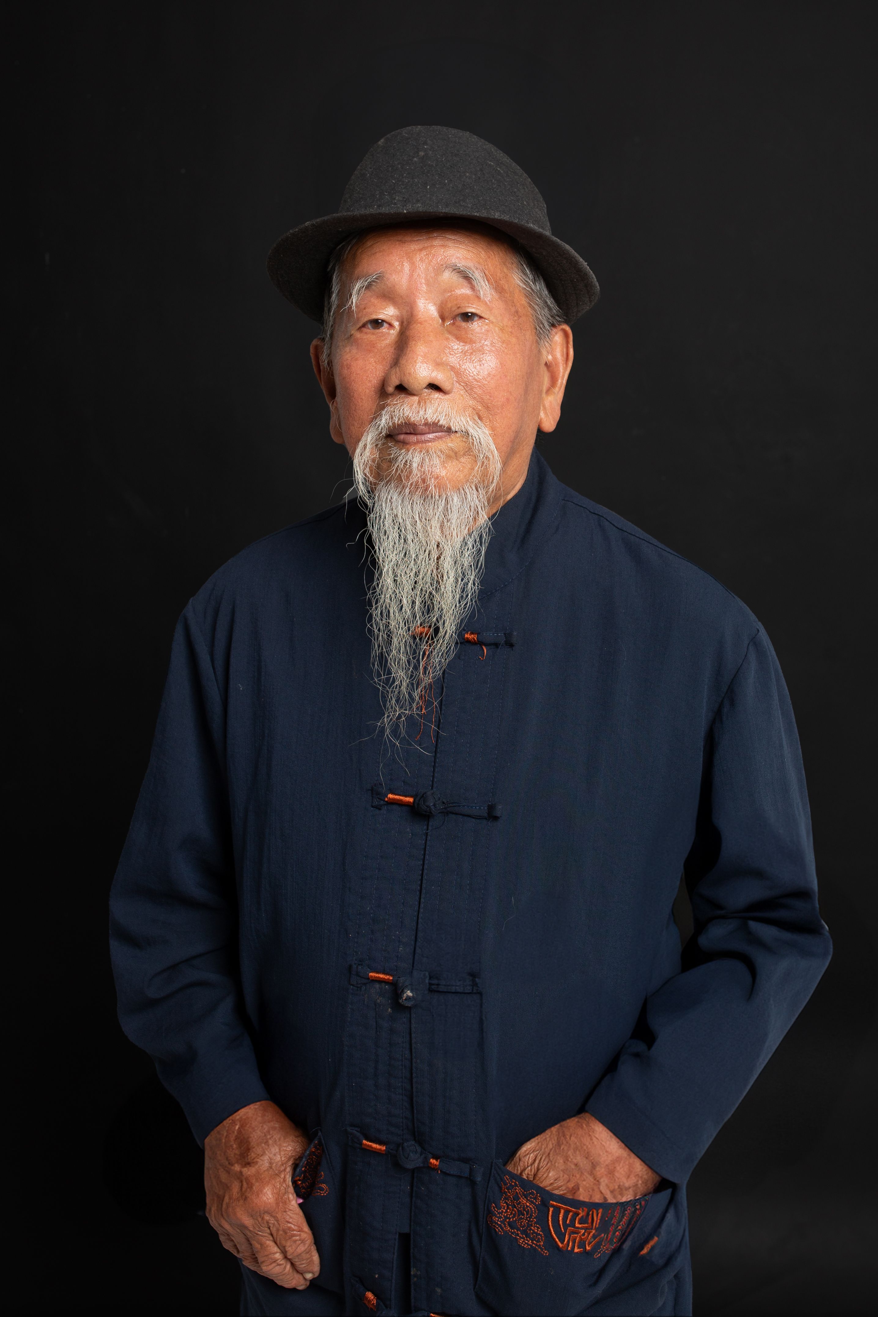 Portrait of an elderly Asian man with a long beard, wearing traditional clothing and a hat, against a dark background.