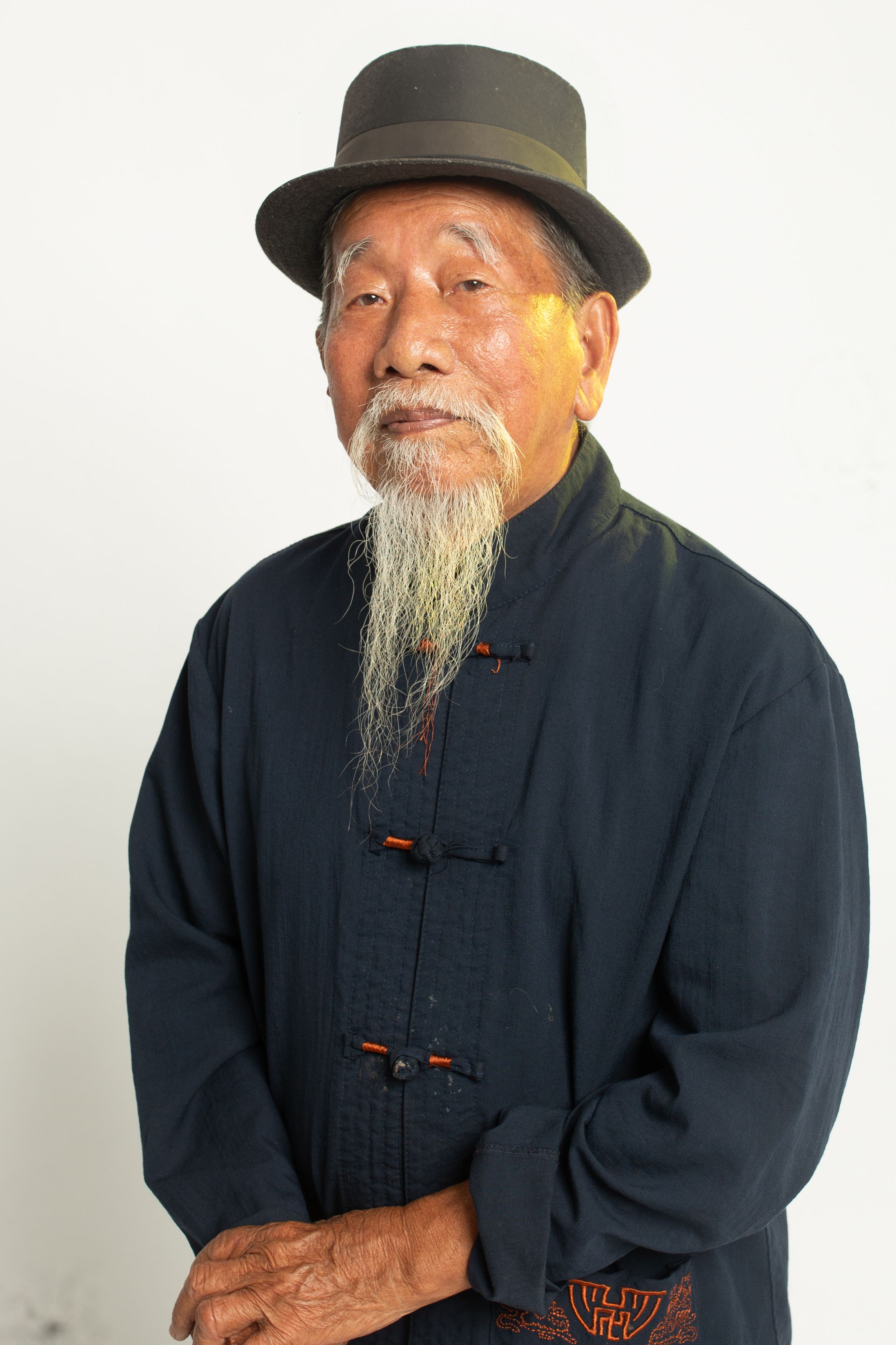 Elderly man with a long beard and hat in traditional attire posing for a studio portrait.