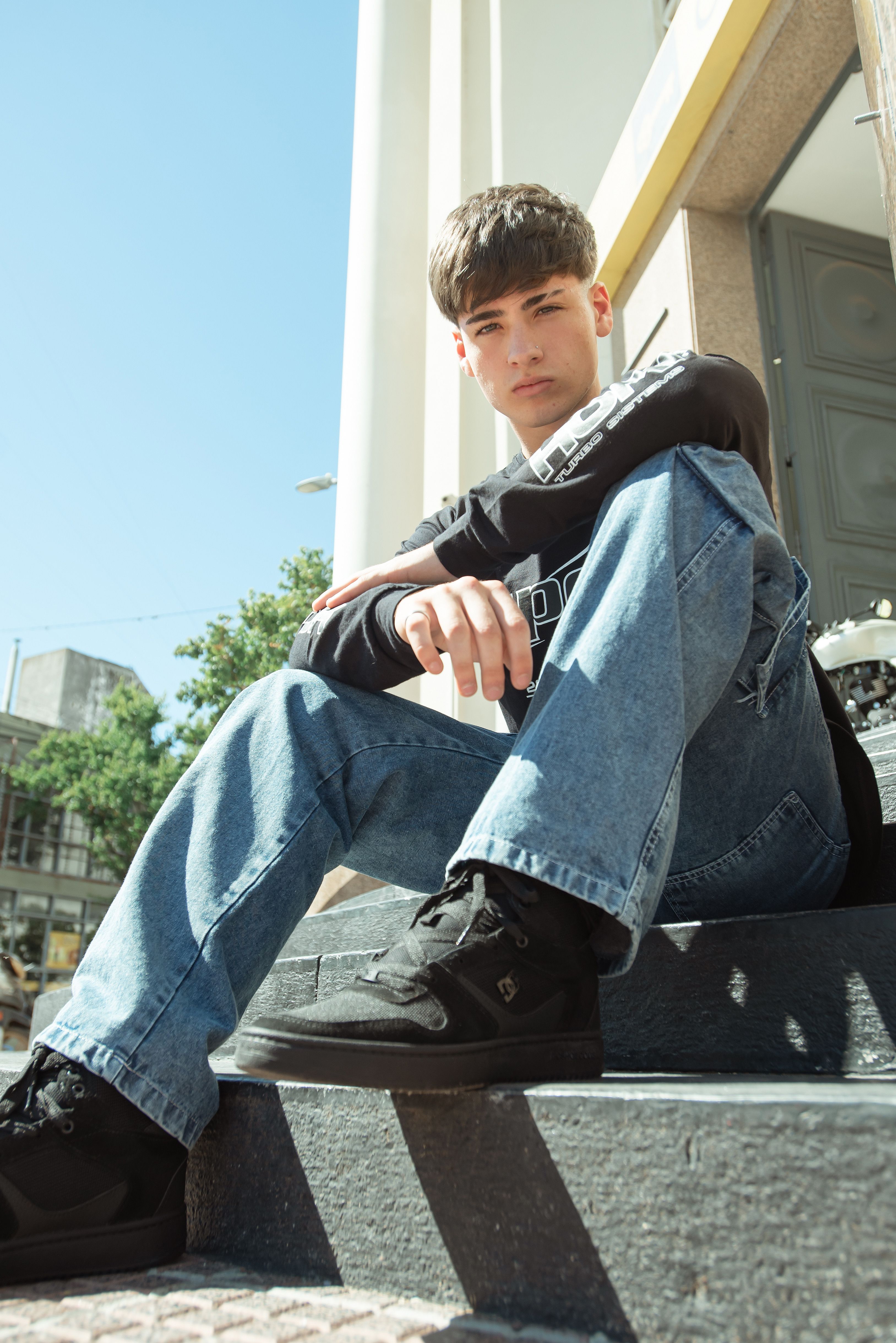 Young man sitting on outdoor steps, posing in jeans and sweatshirt on a sunny day.