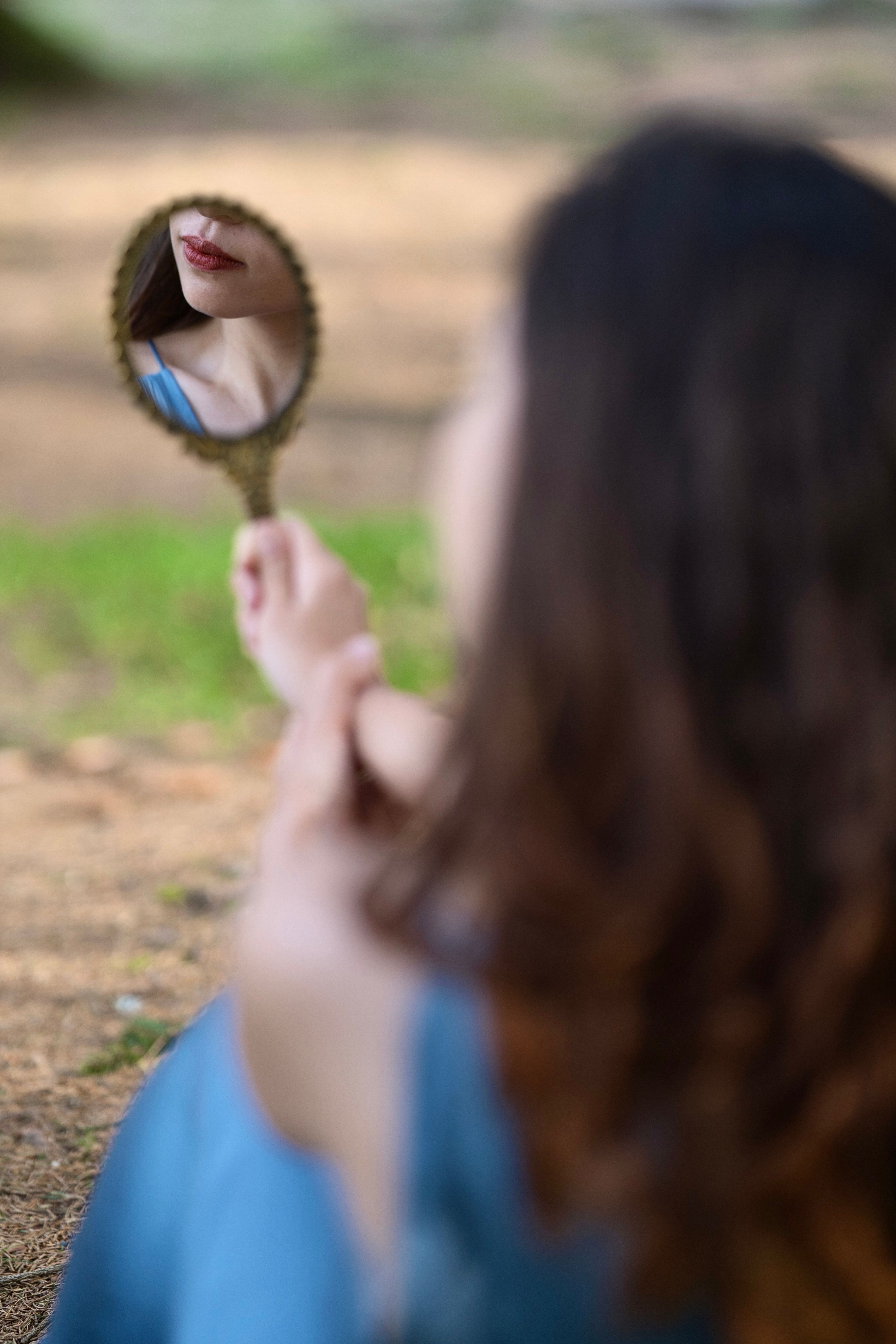 A woman views her reflection in a hand mirror with a blurred natural background, capturing an intimate moment.