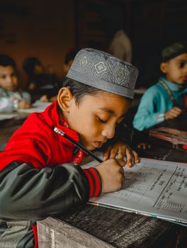 Young boy in Bangladesh concentrates on writing at desk in classroom.
