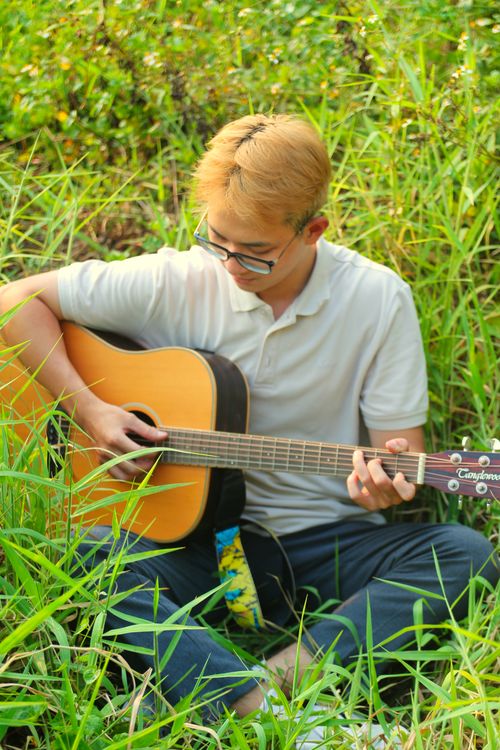 Free A young man sits outdoors, playing an acoustic guitar in a lush, grassy setting. Stock Photo