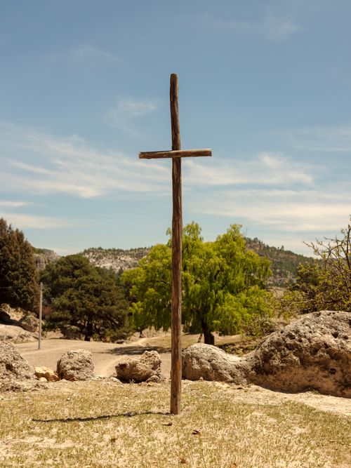 Free A solitary wooden cross stands outdoors in a rural landscape, surrounded by rocks and trees under a blue sky. Stock Photo