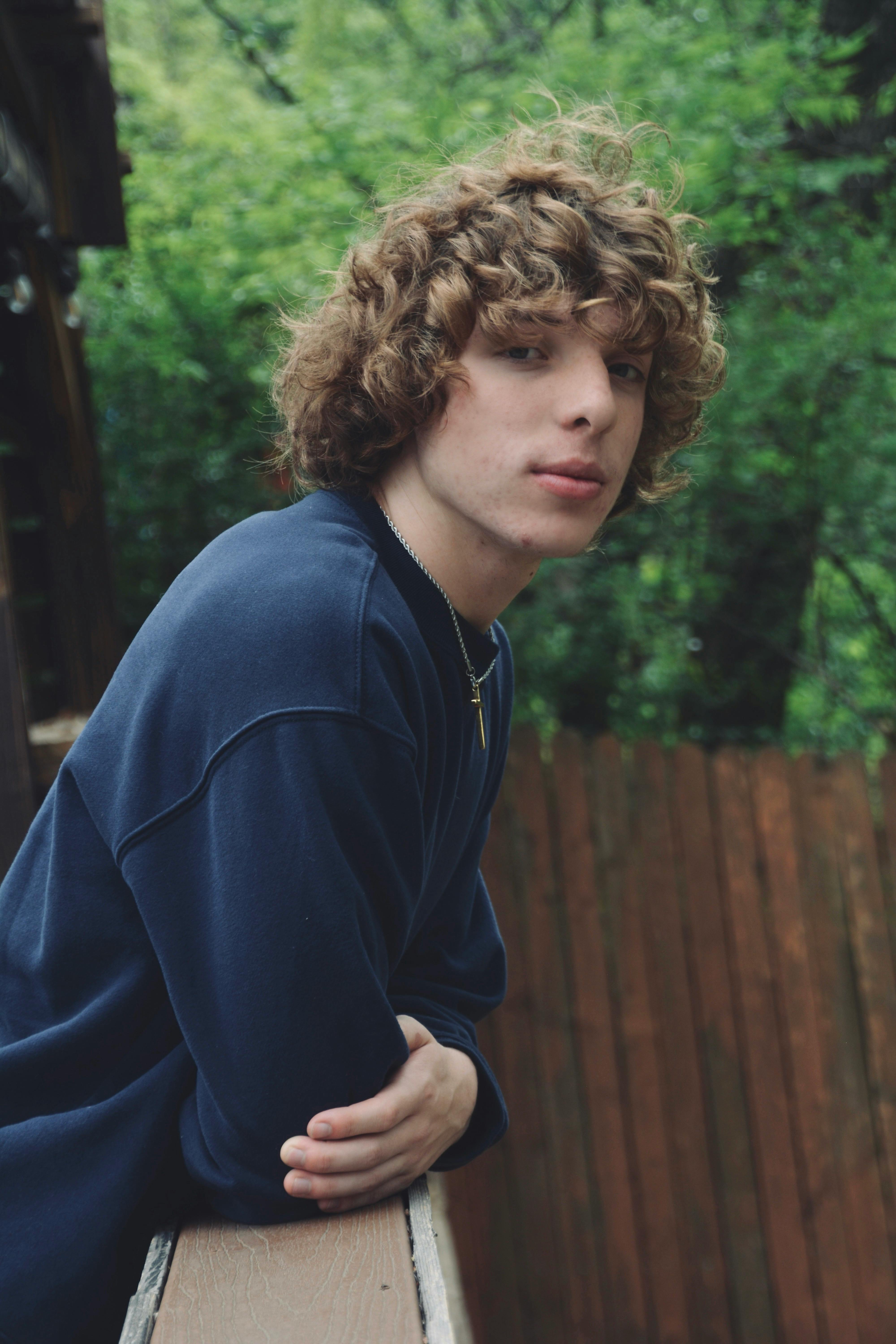 Portrait of a young man with curly hair leaning on a wooden railing with greenery in the background.