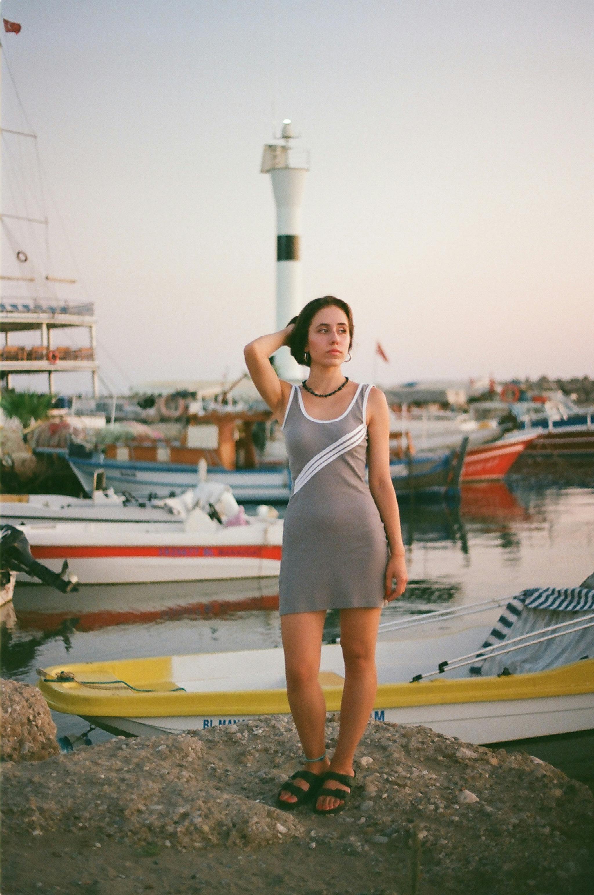 A woman in a stylish dress standing near boats with a lighthouse in the background during sunset.