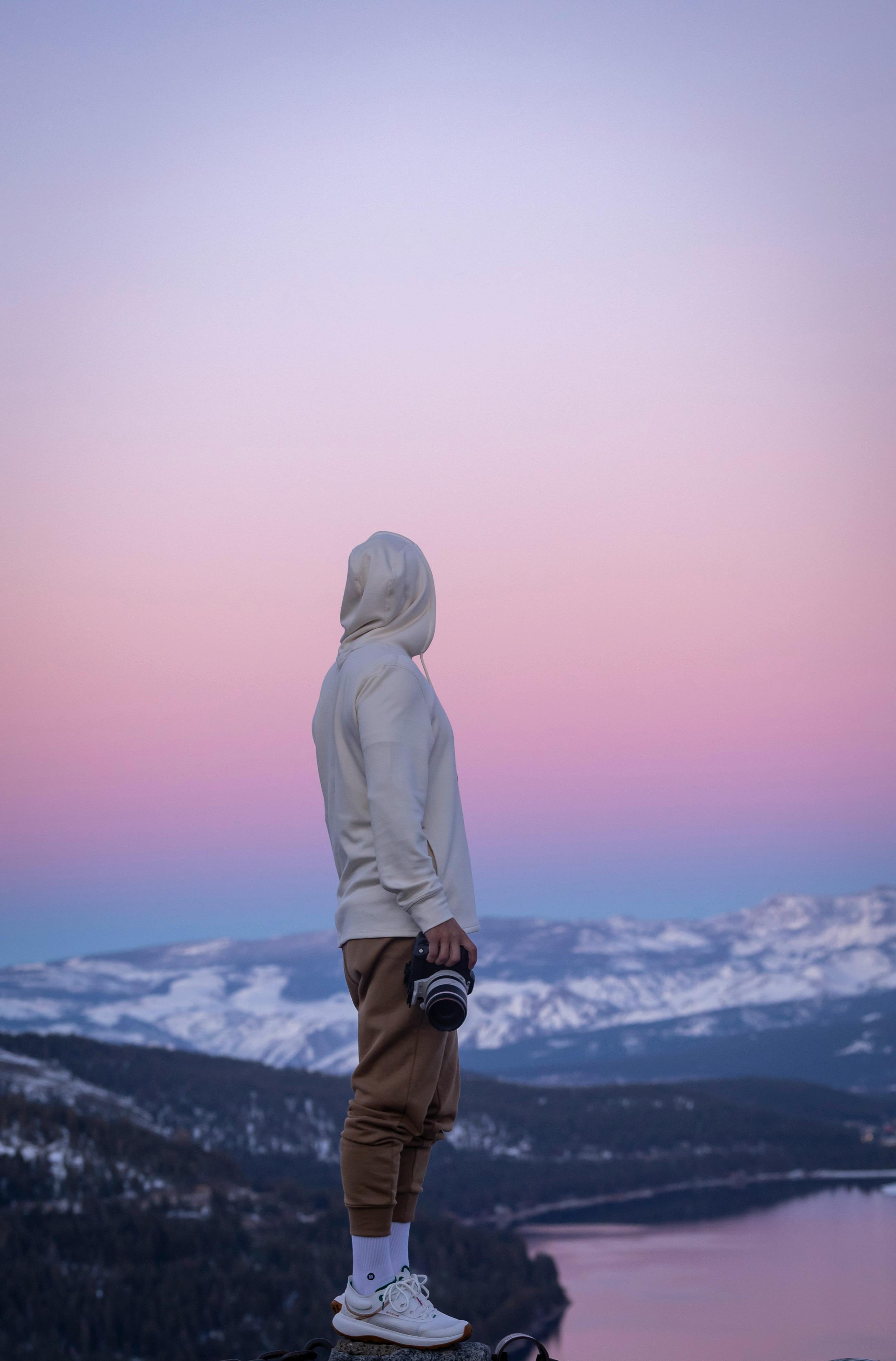 A person in a hoodie stands on a cliff at twilight, overlooking a mountain lake and snow-covered peaks.