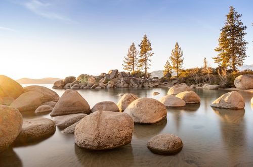 Free Peaceful scene of Lake Tahoe at sunset with rocks and trees reflecting in the calm waters. Stock Photo