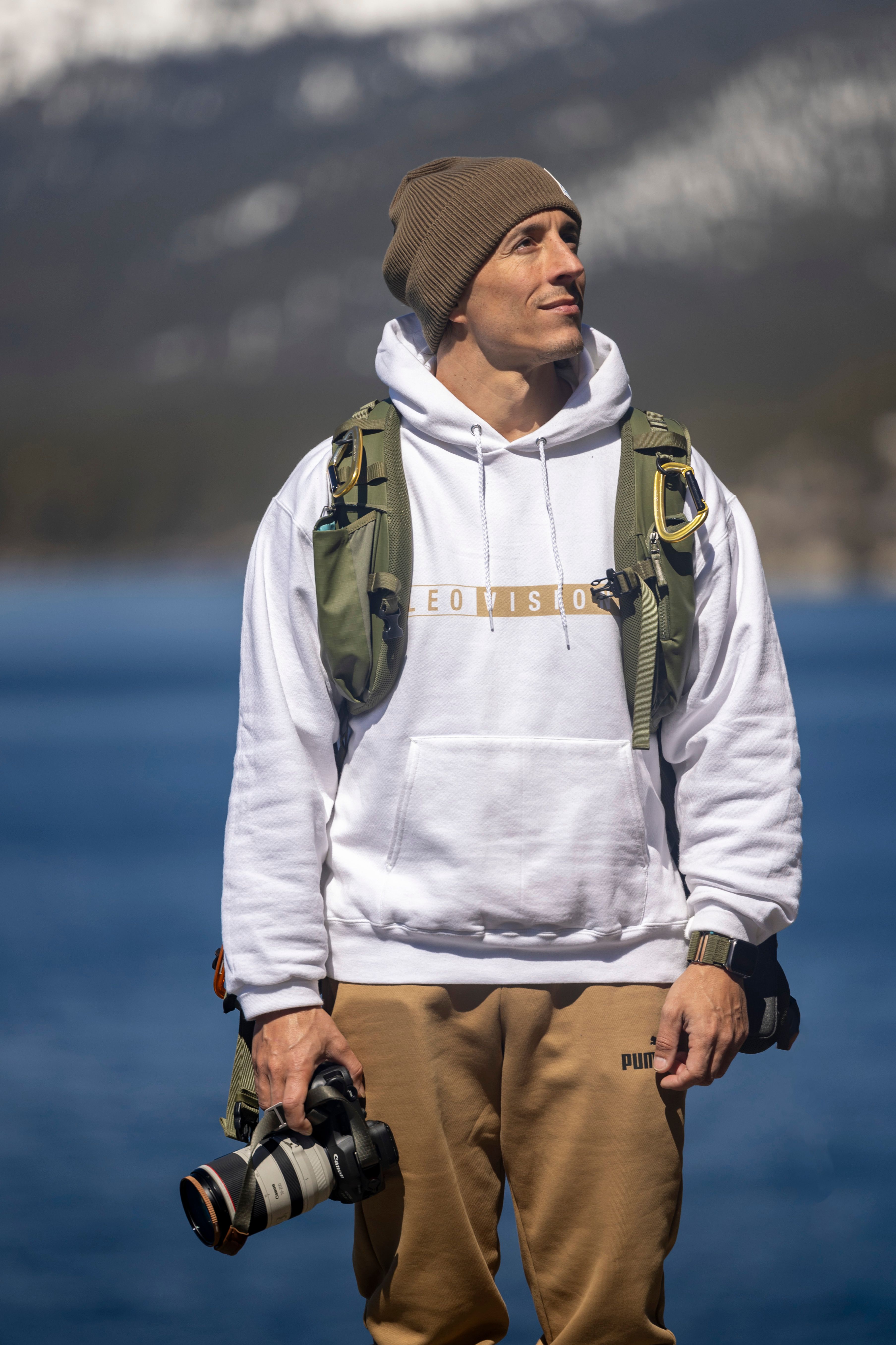 Man in hoodie with camera stands by lake on a sunny day