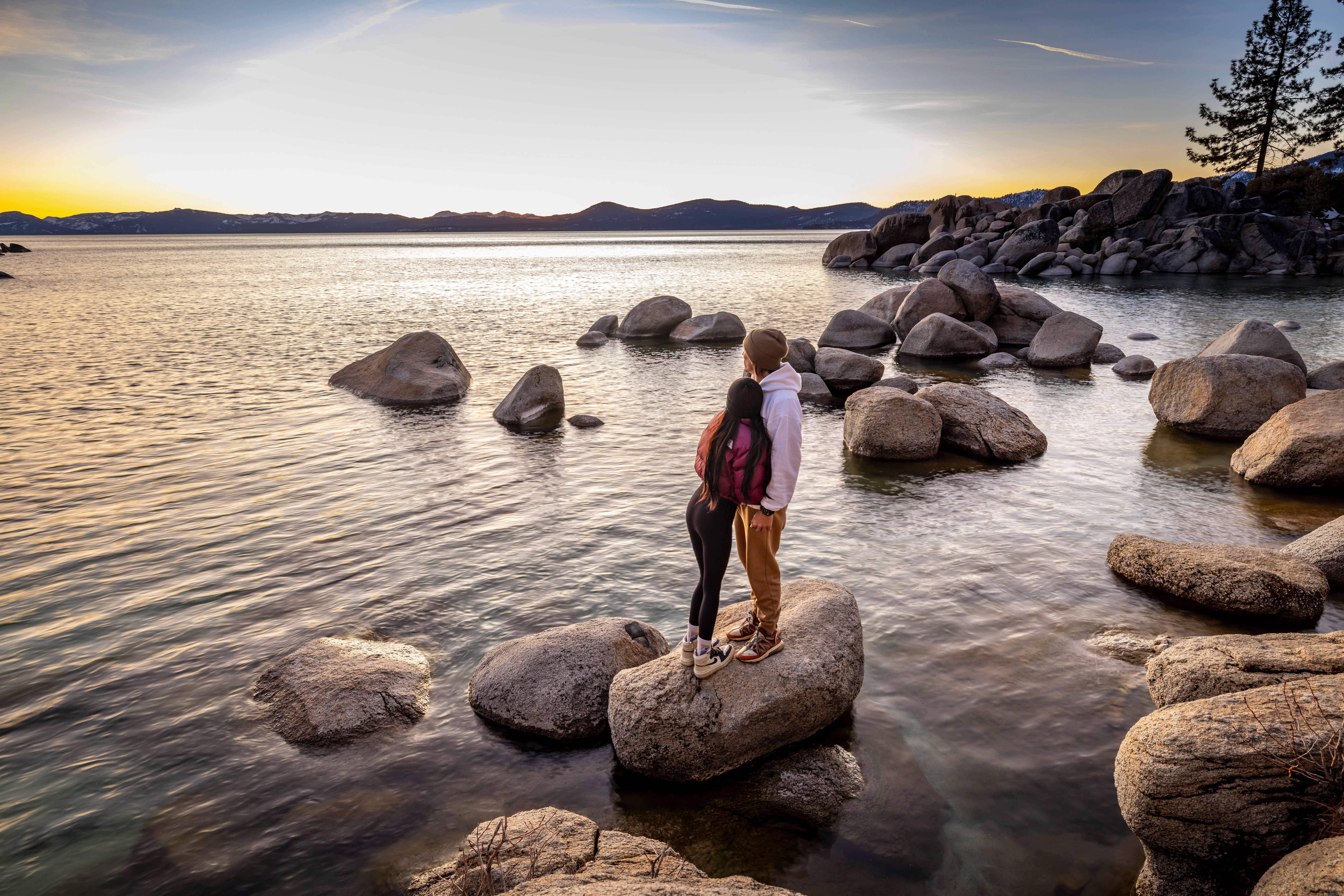 Young couple stands on rocks by a peaceful lake at sunset, embracing tranquility.