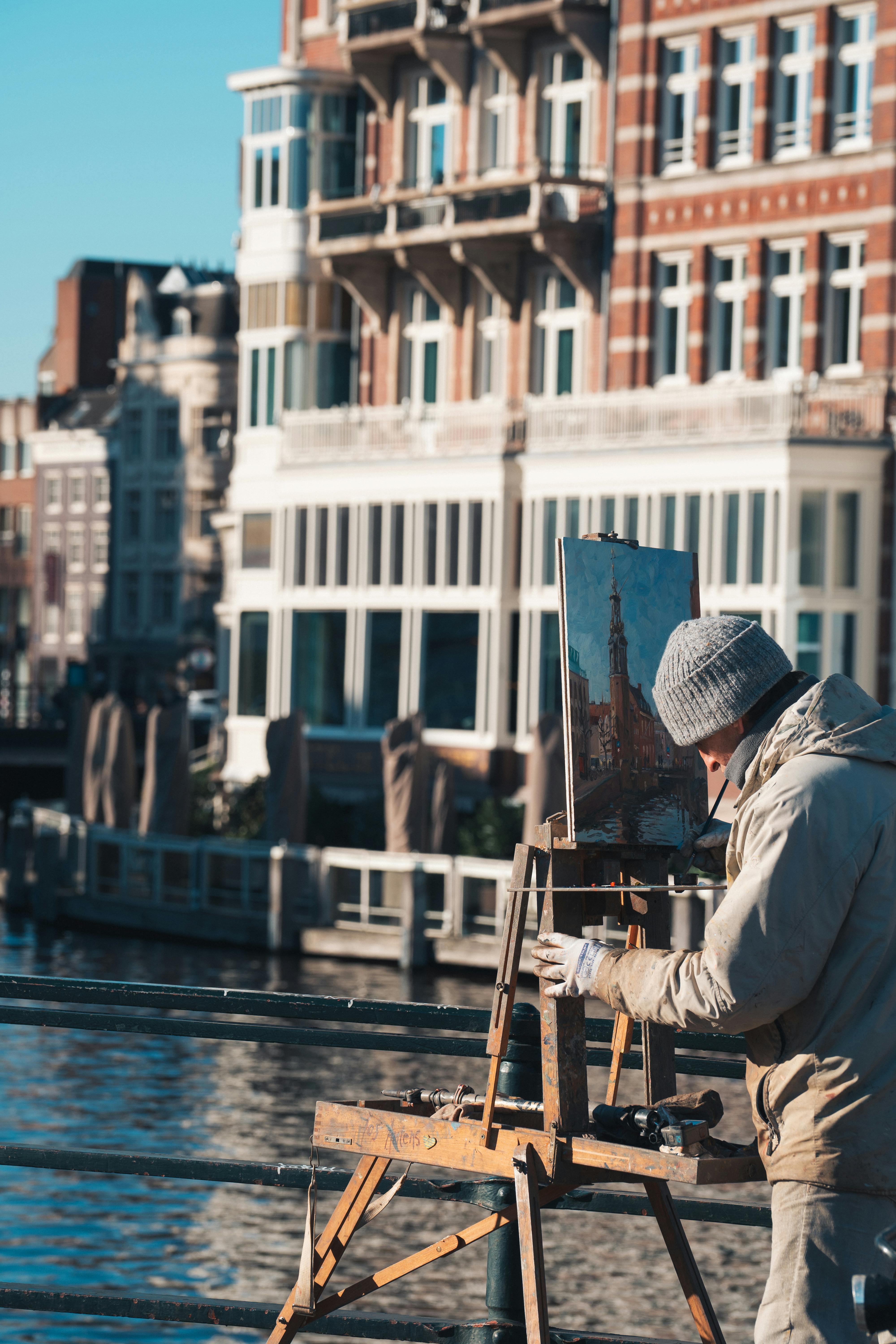 An artist paints outdoors along a sunny canal with historic buildings in Amsterdam.