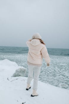A woman stands by the snowy coast wearing a warm winter jacket.