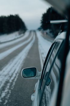 A scenic view from a car on a snowy road, highlighting the rear-view mirror in a winter setting.
