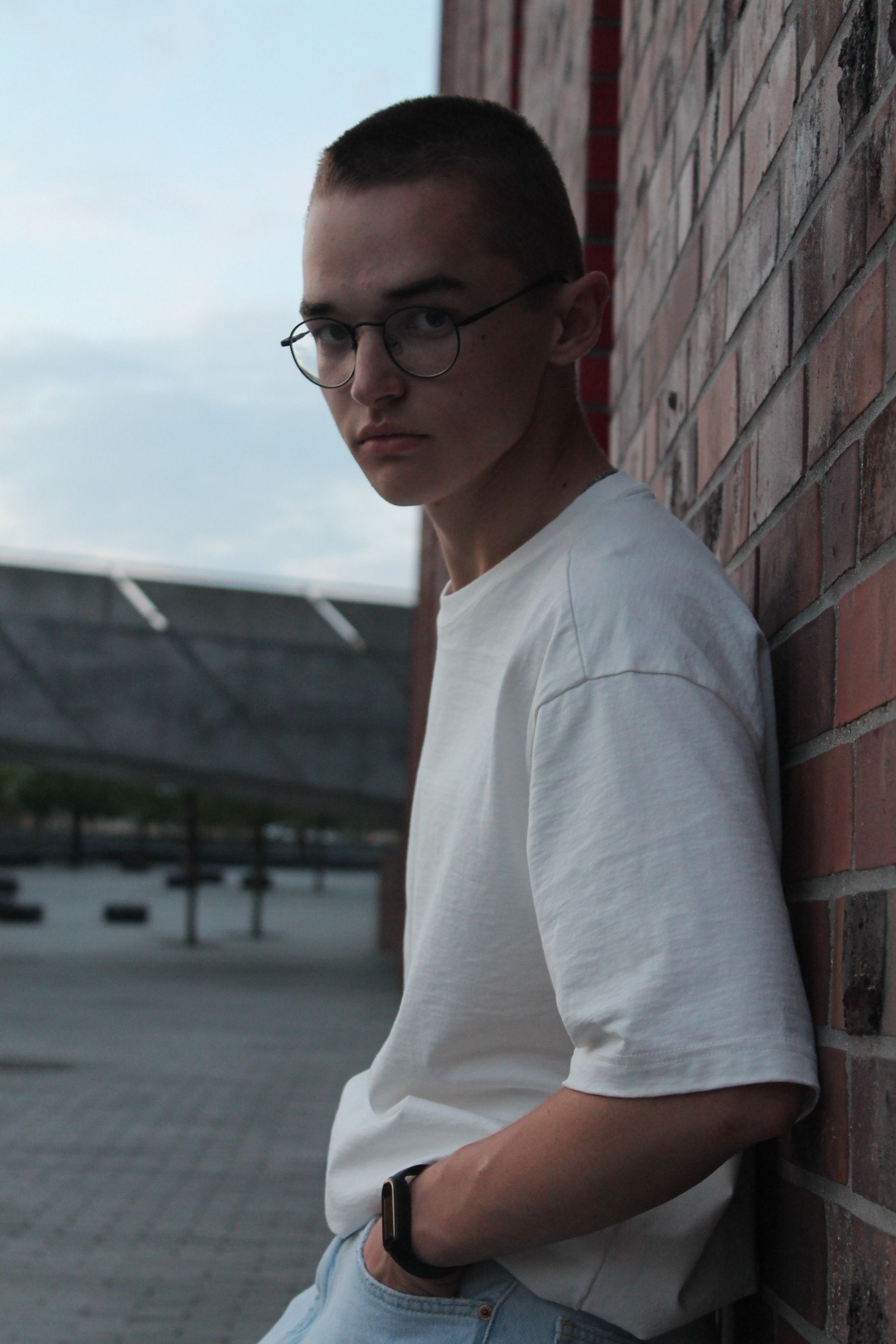 Portrait of a young man with glasses leaning against a brick wall in a city landscape.