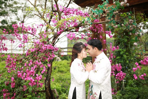 A couple in traditional clothing pose for a photo in front of a flowering tree