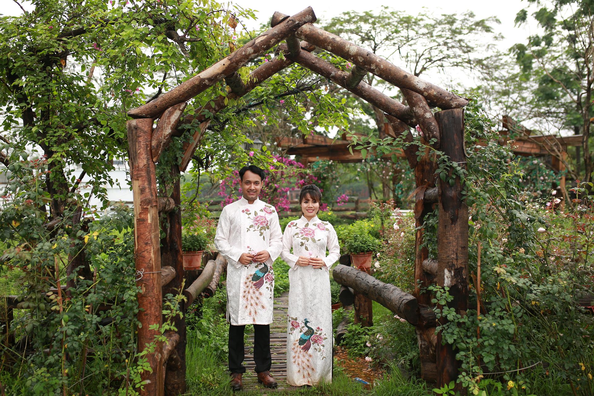 Happy couple in traditional clothing standing under a wooden pergola in a lush garden.