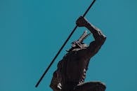 在晴朗的蓝天下，以低角度拍摄了一座手持长矛、姿态动态的土著战士雕像